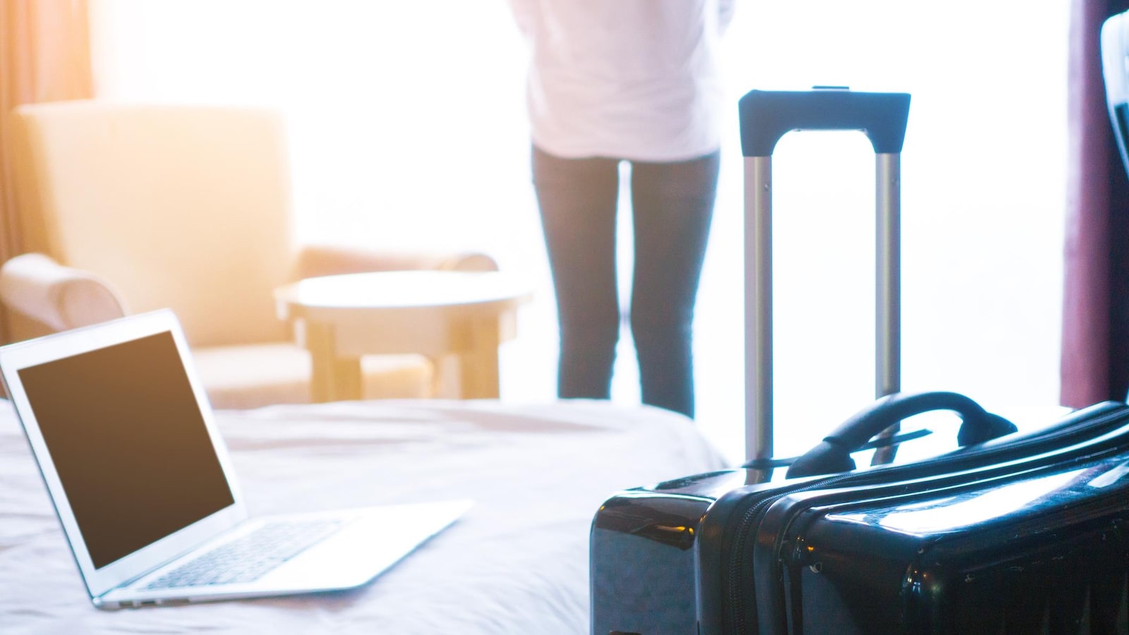 Une photo montrant une femme de dos en train de regarder à la fenêtre de son hôtel. En avant-plan, on peut voir une valise et un ordinateur portable posé sur le lit.