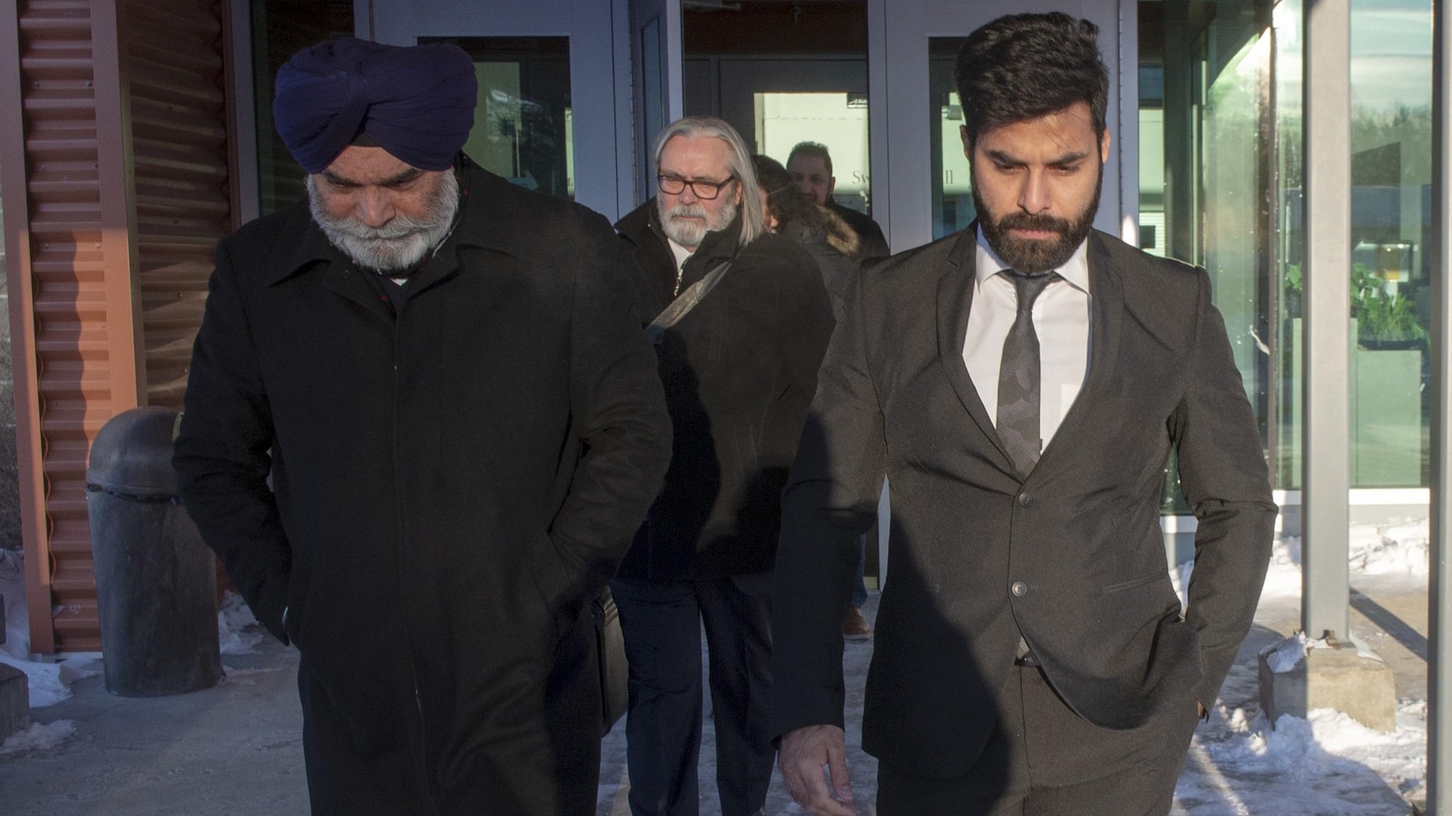 Un jeune homme portant un complet avec une cravate sort à l'extérieur d'un bâtiment accompagné d'un autre homme, barbu et portant un turban.