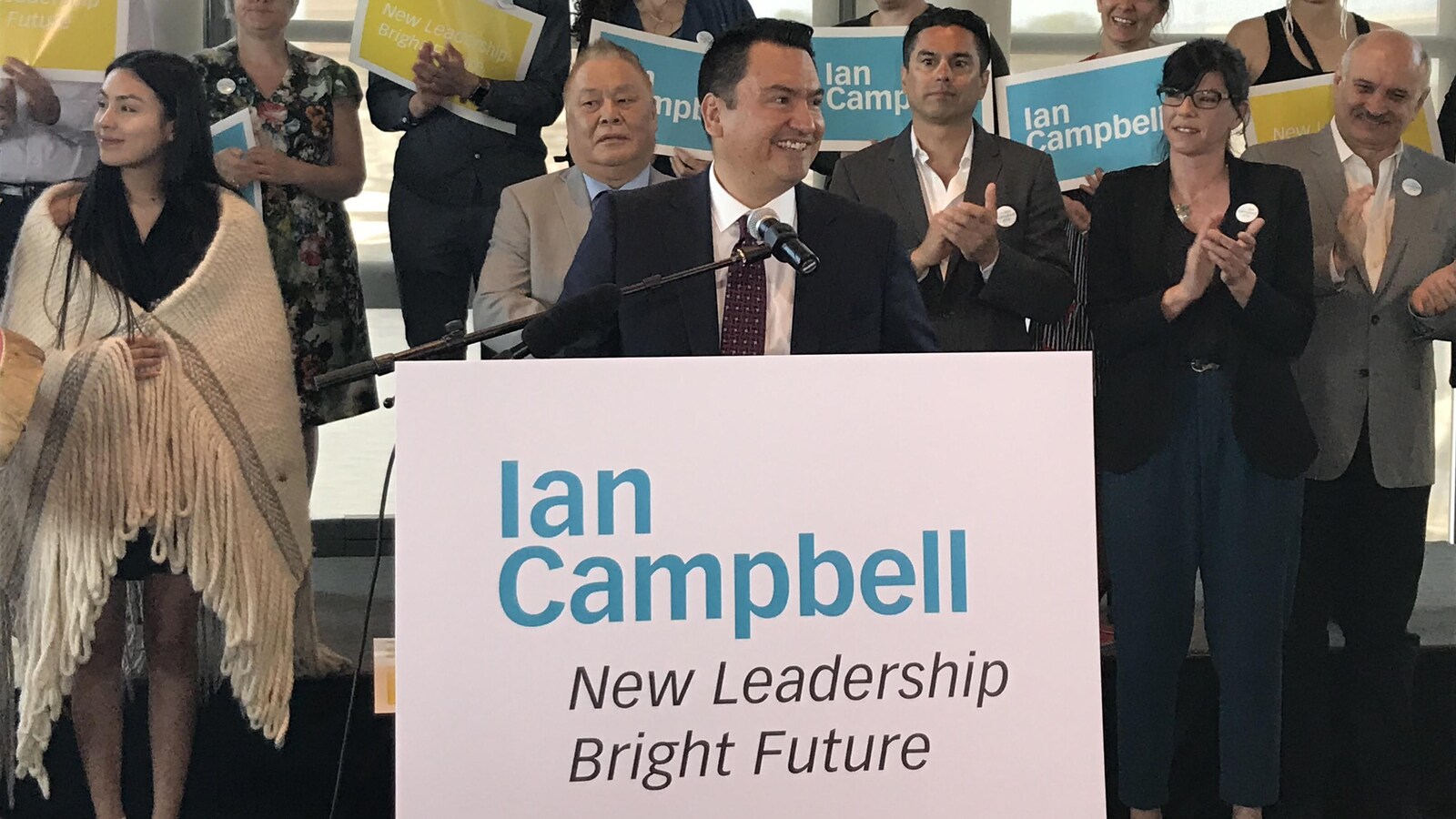 Ian Campbell devant un micro qui sourit. Il porte un complet bleu avec une cravate bourgogne.