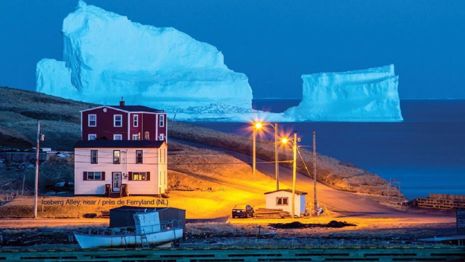Deux maisons illuminées par la lumière jaunâtre des lampadaires, un bateau de pêche accosté sur le rivage et un énorme iceberg bleuté en arrière-plan.