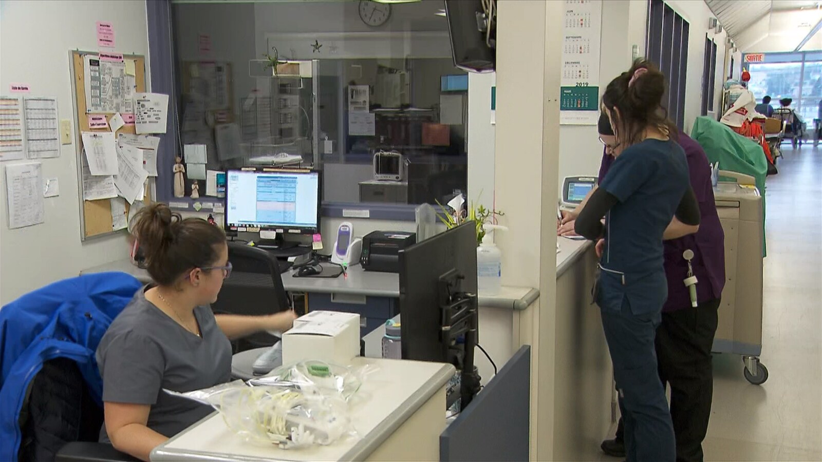 Une infirmière est assise à son poste d'ordinateur et deux autres debout à remplir des formulaires.