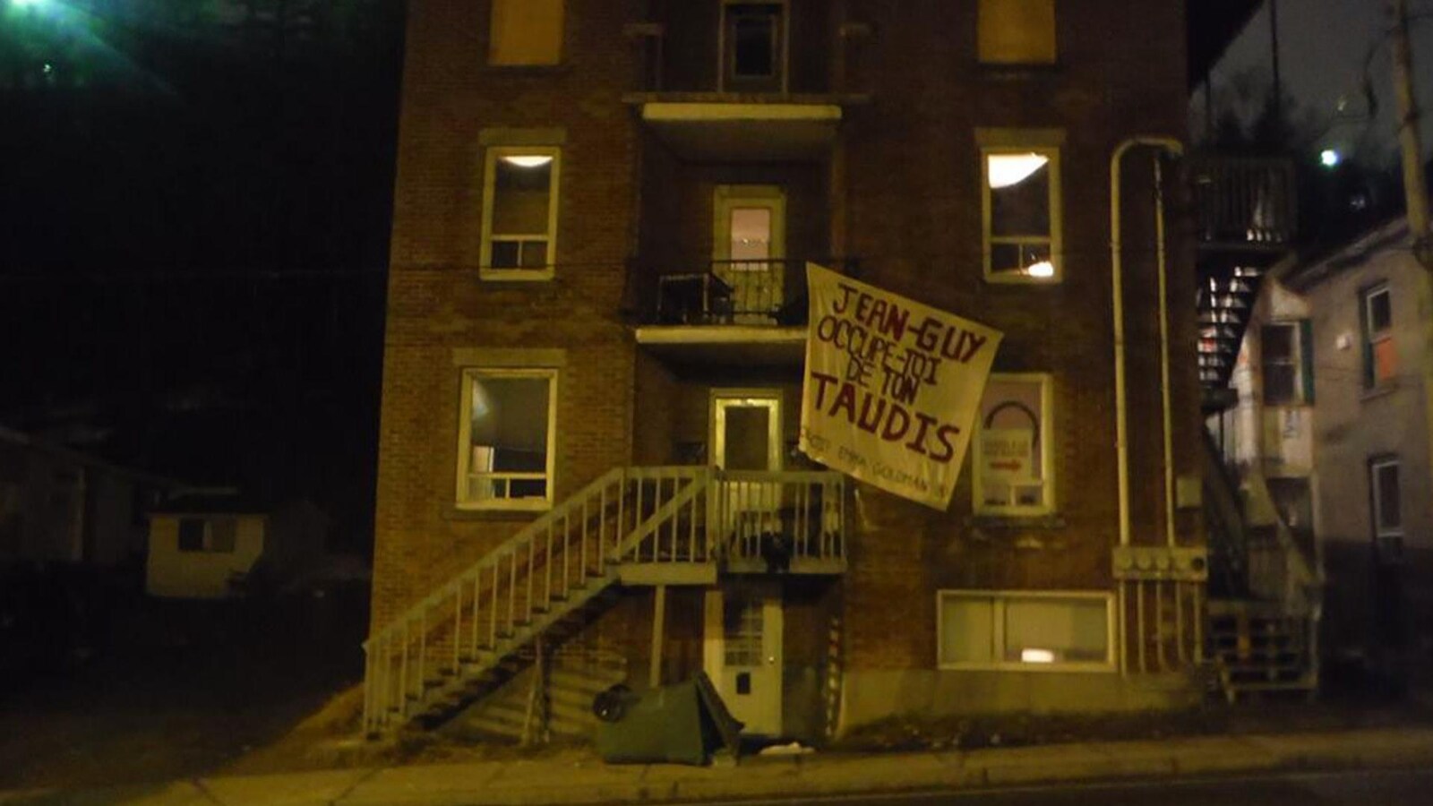 Une affiche qui est inscrit : Jean-Guy, occupe-toi de ton taudis est accroché sur l'immeuble d'un logement.