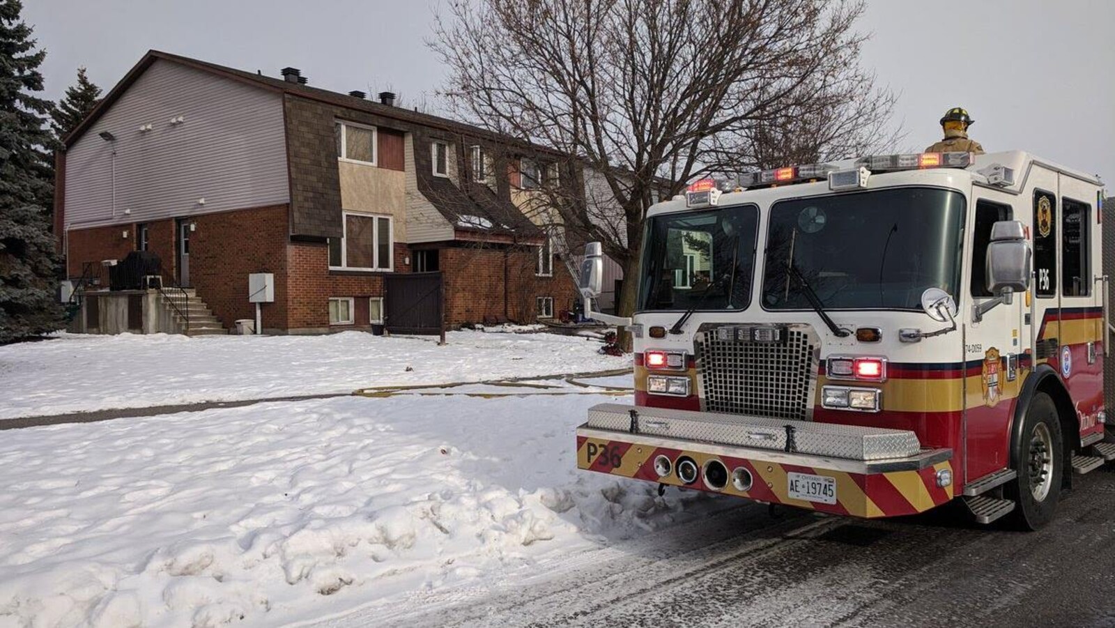 Un camion de pompiers avec les gyrophares allumés devant un immeuble à logements et un terrain enneigé.