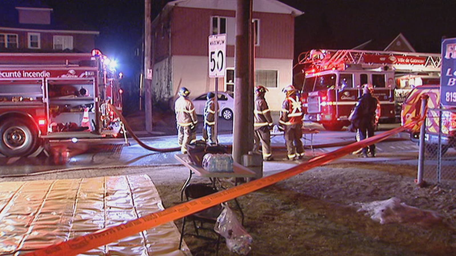Des pompiers en uniforme derrière un périmètre de sécurité. Dans la rue, des camions avec leurs gyrophares allumés.