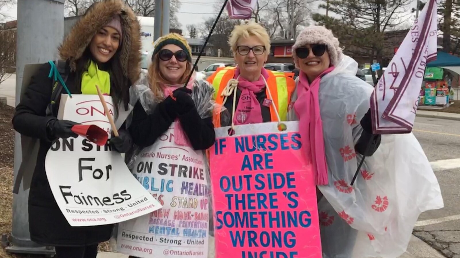 quatre femmes portant des vêtement chauds et colorés se tiennent l'une à côté de l'autre, elles portent des bannières et pancartes aux messages de solidarité pour les infirmières en grève, dont elles font partie.