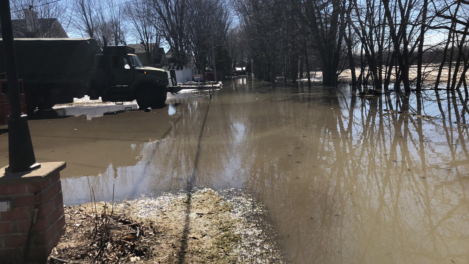 Dans un secteur rural, de l'Eau inonde ce qui était une rue. Un camion de l'armée est stationné devant des maisons.