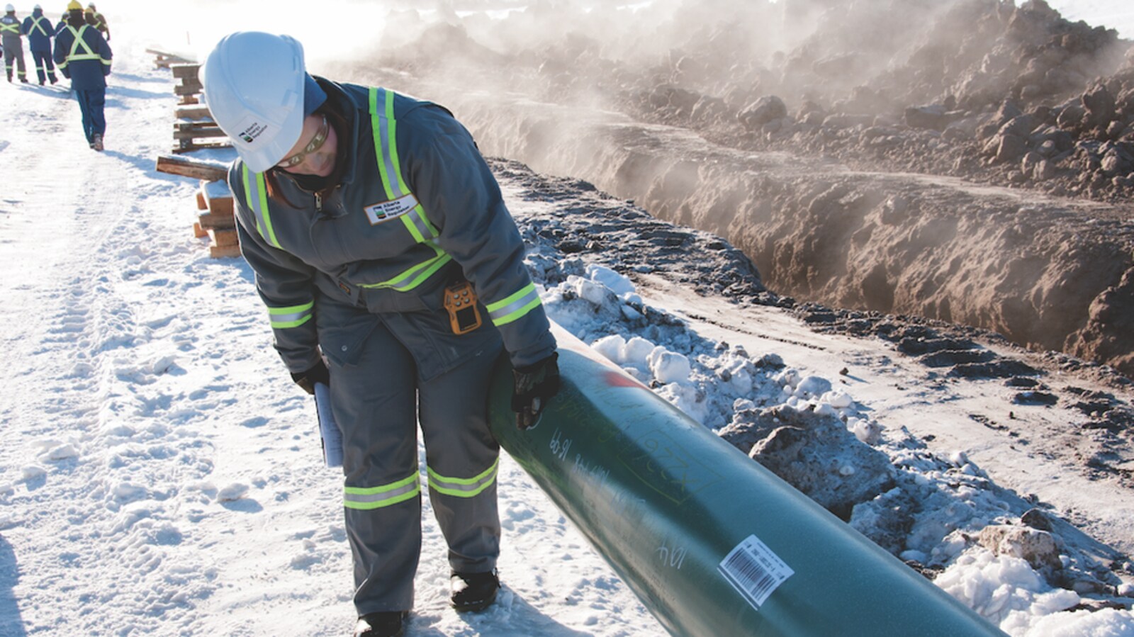 Une personne portant un casque de sécurité passe une main gantée sur un pipeline sur un chantier enneigé.