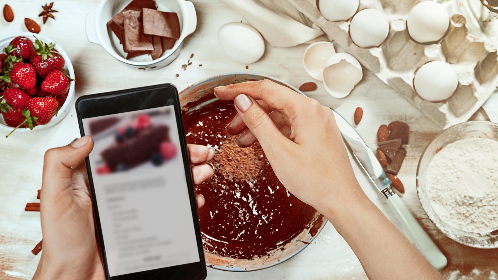 Une personne saupoudre de cacao un gâteau au chocolat en lisant les indications de la recette sur son cellulaire.
