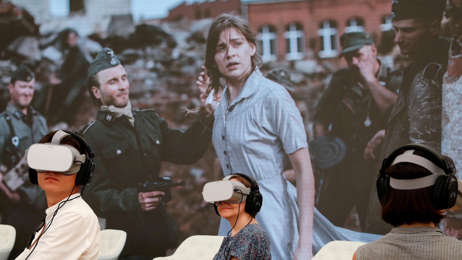 Des personnes portent des casques de réalité virtuelle et des écouteurs devant une murale montrant une jeune femme apeurée entourée de soldats nazis.
