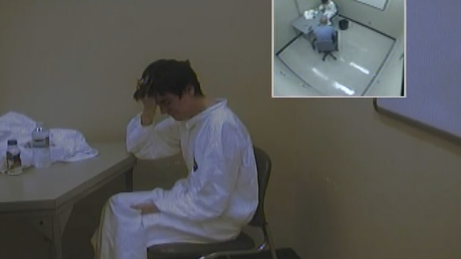 Image d’Alexandre Bissonnette tirée d’une caméra de surveillance. Il est assis sur une chaise, face à une table. Vêtu d’une combinaison blanche, il se prend la tête avec sa main droite. Dans le coin supérieur droit de l’image, on aperçoit une autre prise de vue de la caméra, en plongée. Le sergent enquêteur Steve Girard, de la Sûreté du Québec, fait face à Bissonnette.