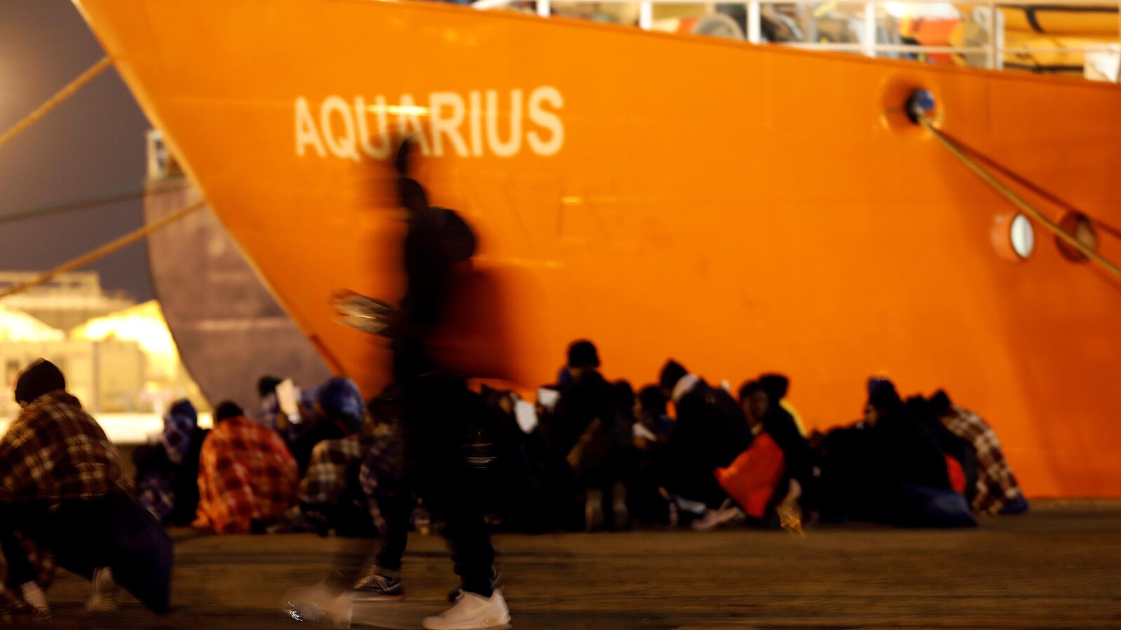 Des gens sont assis dans un port, un énorme bateau orange est amarré près d'eux.