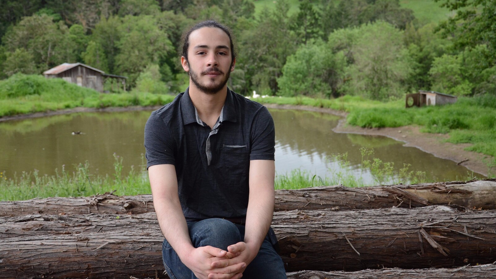 Jacob Lebel, assis sur un tronc d'arbre, devant une étendue d'eau et un boisé.