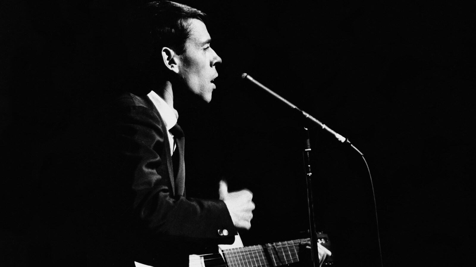 Sur une scène, Jacques Brel donne une performance s'accompagnant de sa guitare, un micro sur pied devant lui.