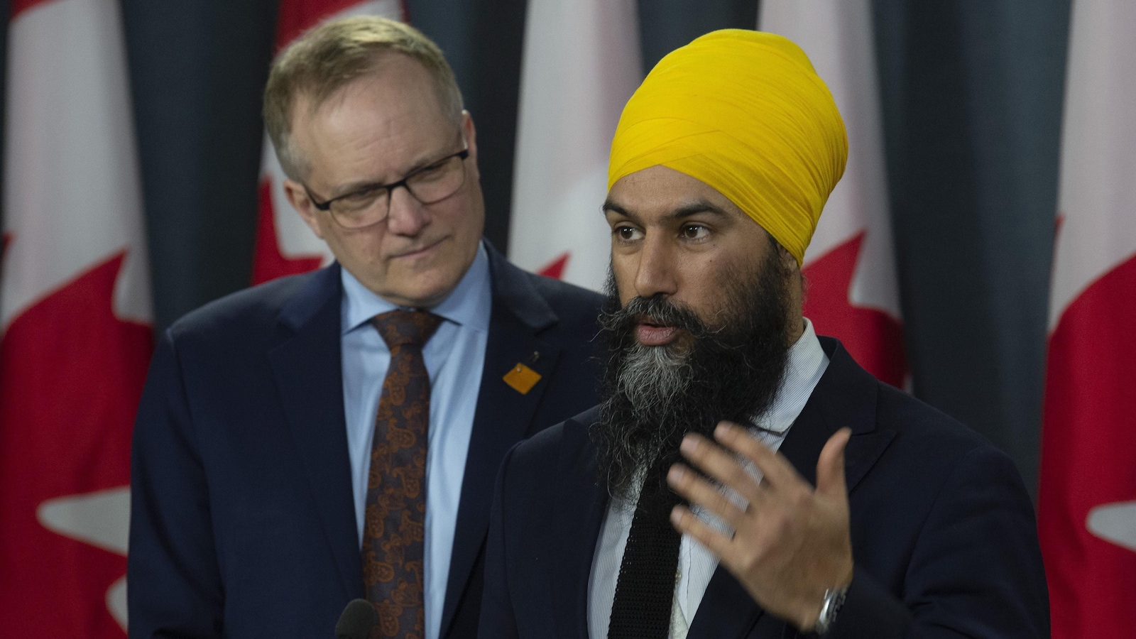 Une homme portant la barbe longue et un turban s'adresse au micro. Derrière lui, une rangée de drapeaux du Canada. Un homme en veston se tient à ses côtés.