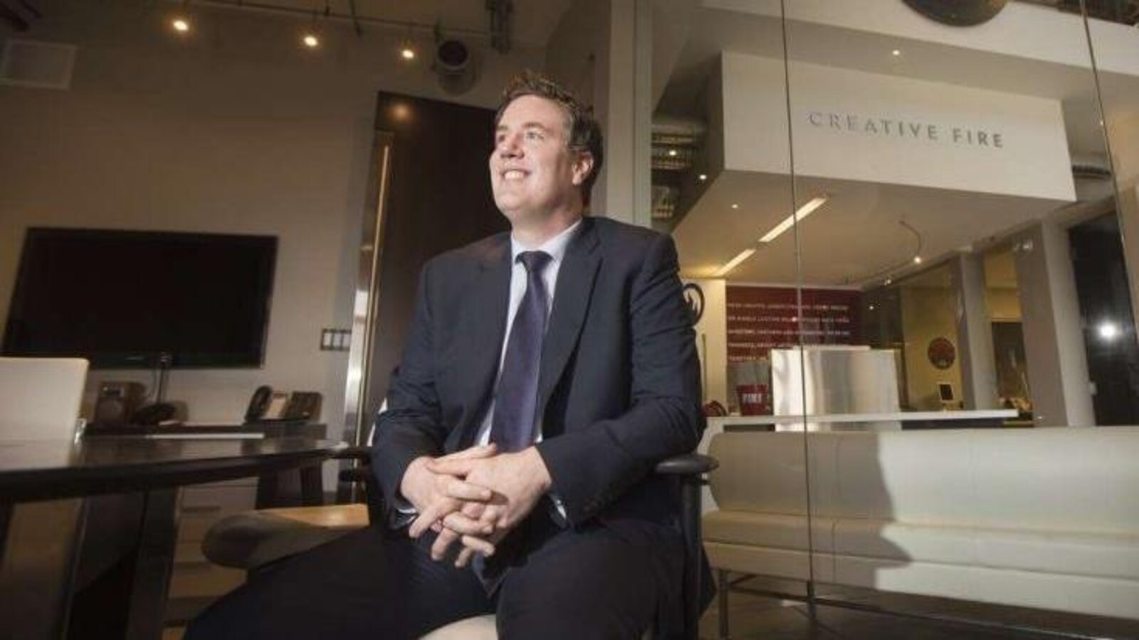 Un homme photographié en contre-plongée, en costume cravate et assis dans un bureau au design moderne, sourit en regardant vers une lumière devant lui. Derrière lui se trouve une baie vitrée donnant sur l'intérieur des locaux de l'entreprise Creative Fire.