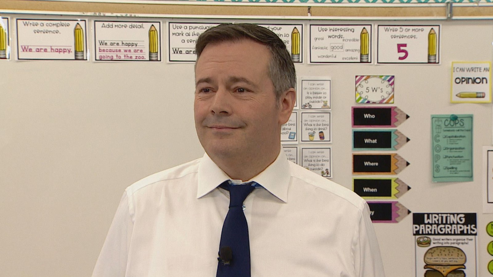Jason Kenney sourit devant un tableau dans une classe.