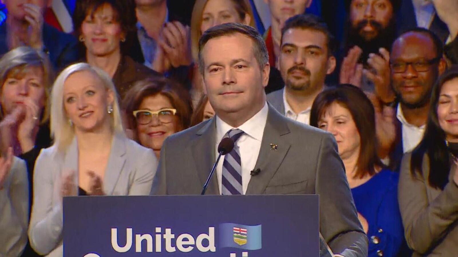 Avec de nombreux supporteurs derrière lui, Jason Kenney qui porte un costume-cravate se tient devant un podium.
