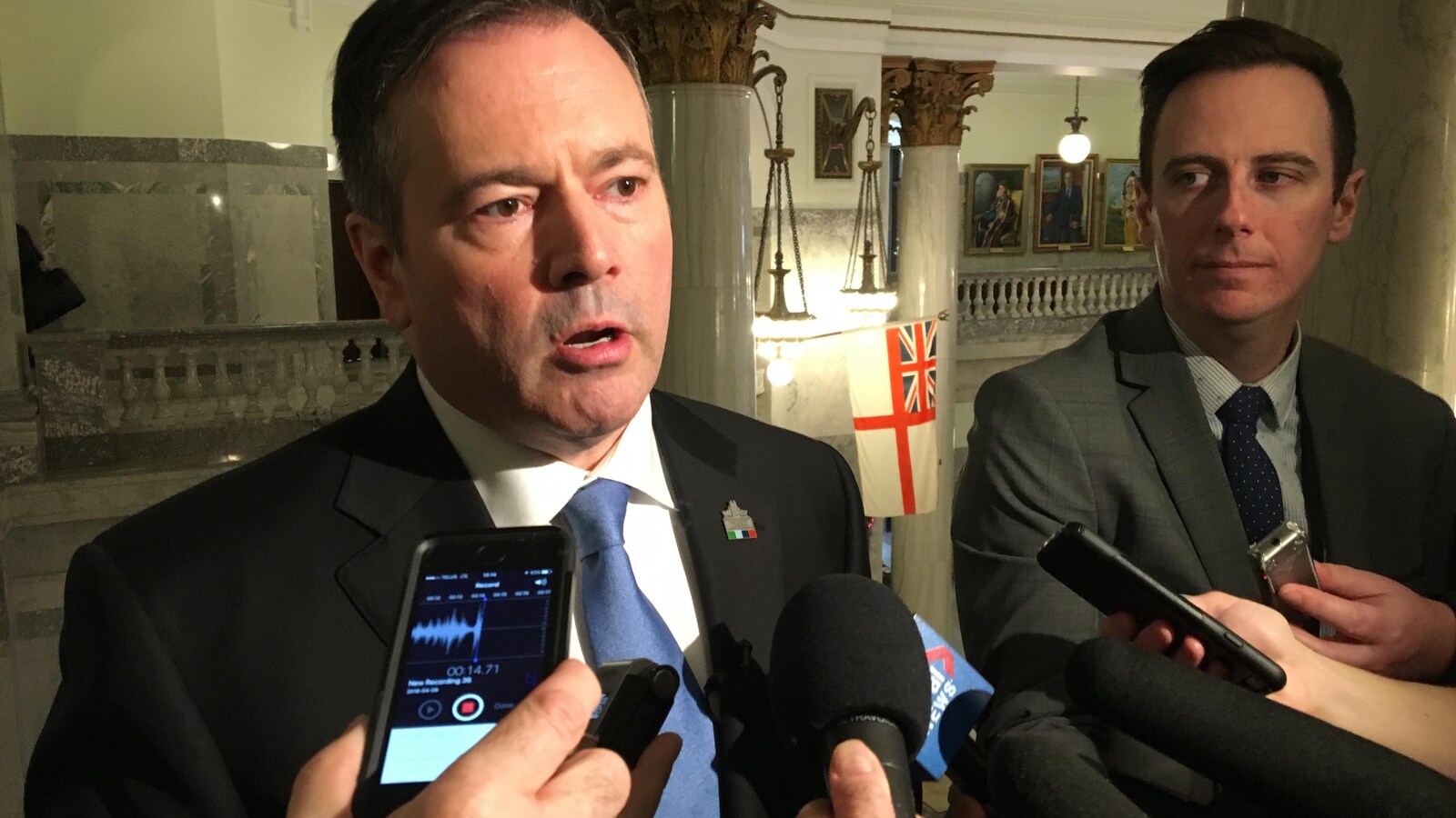 Jason Kenney est de face, il porte une cravate bleue et un veston. En arrière-plan, les colonnes de marbres de l'Assemblée législative de l'Alberta.