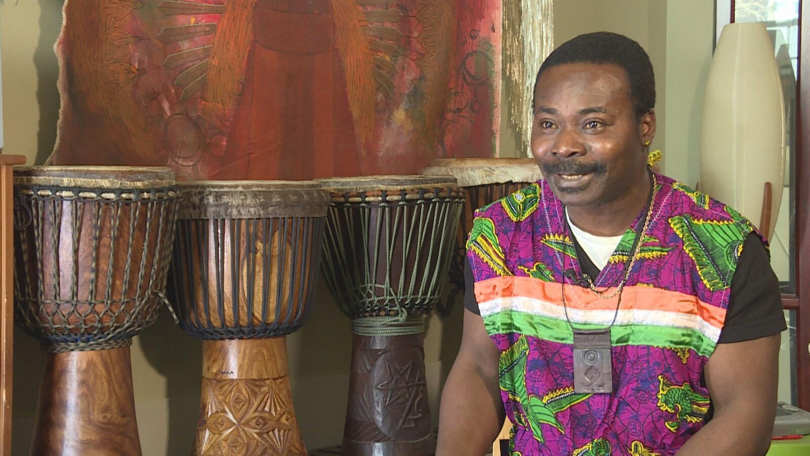 Jean avec porte un vêtement traditionnel de Côte d’Ivoire. Derrière lui, se trouvent 4 grands tambours.