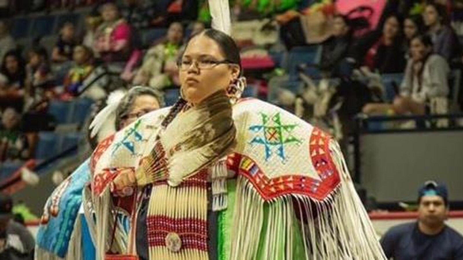 Une jeune femme, debout, dans son costume de danse traditionnel coloré, lors d'une compétition de pow-wow.