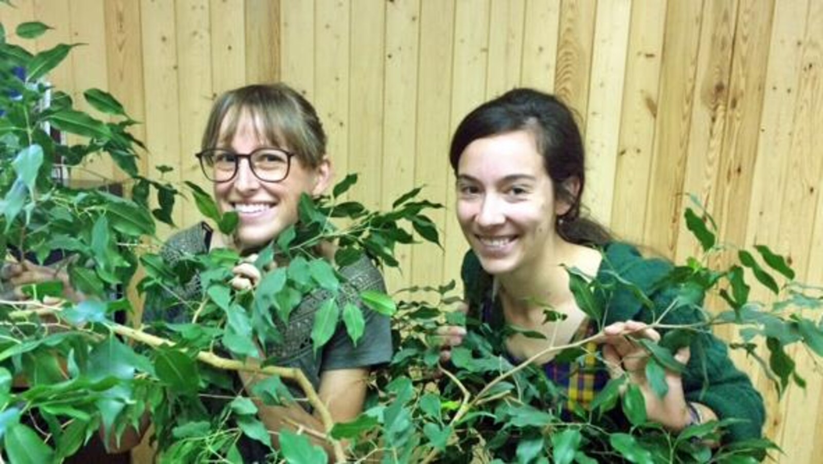 Deux jeunes femmes sourient à la caméra, cachées derrière un petit arbre.