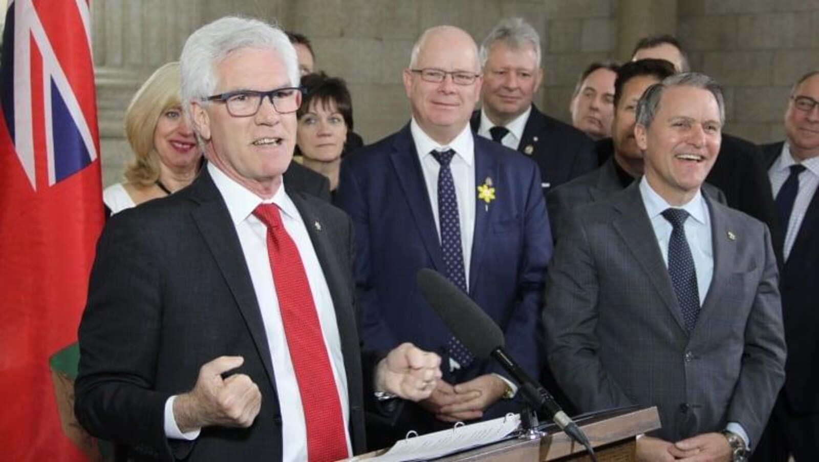 Le ministre Jim Carr, portant une cravate rouge, parle au podium devant une dizaine de personnes et le drapeau manitobain.