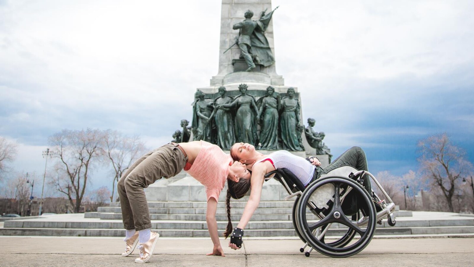 Les danseuses Joannie Douville et France Geoffroy (à droite, en fauteuil roulant) se penchent en arrière pour une chorégraphie effectuée à côté d'une colonne où se trouvent plusieurs statues.