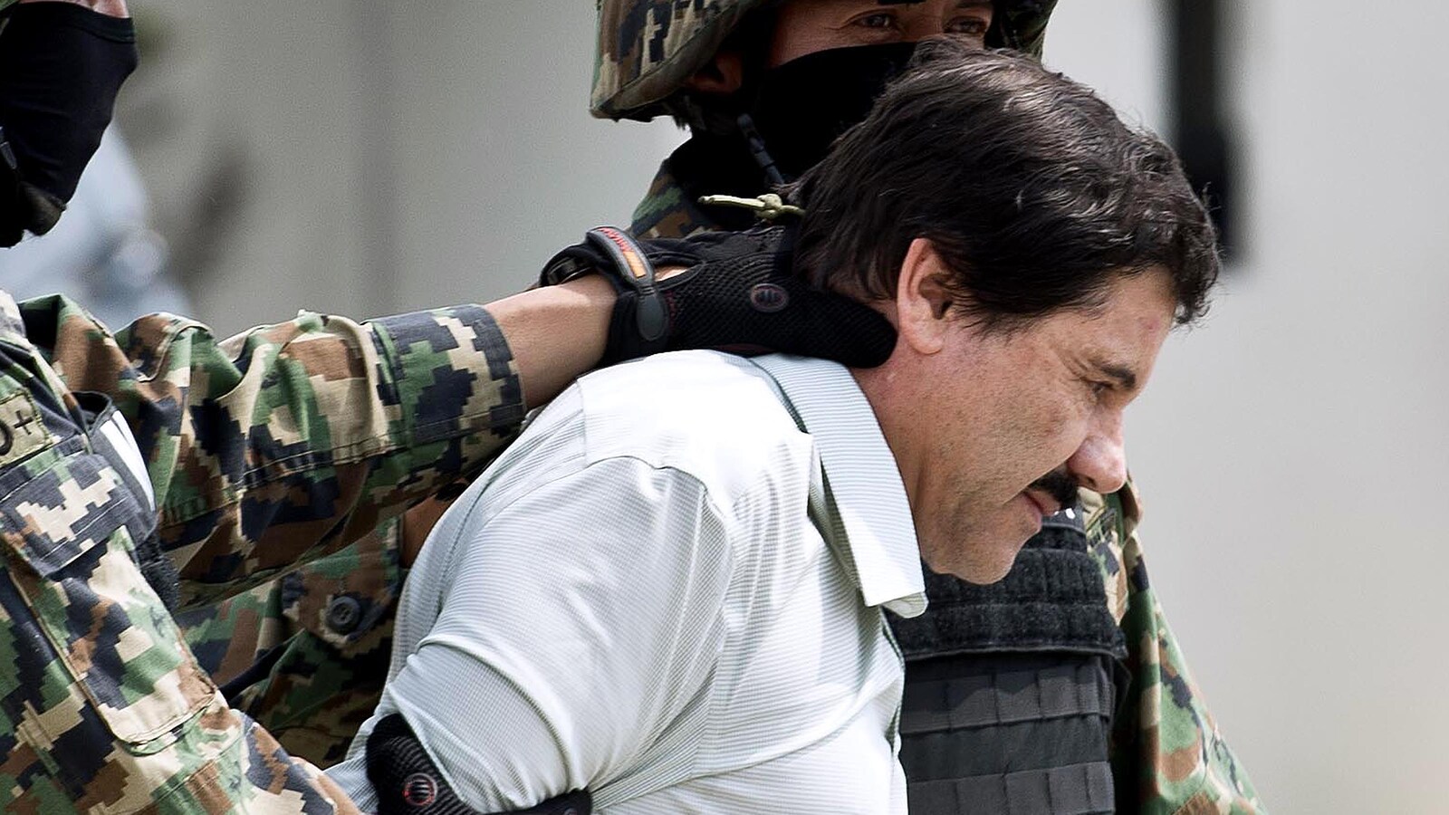 Deux soldats flanquent Joaquin El Chapo Guzman qui apparait en chemise blanche, la tête légèrement penchée vers le bas.