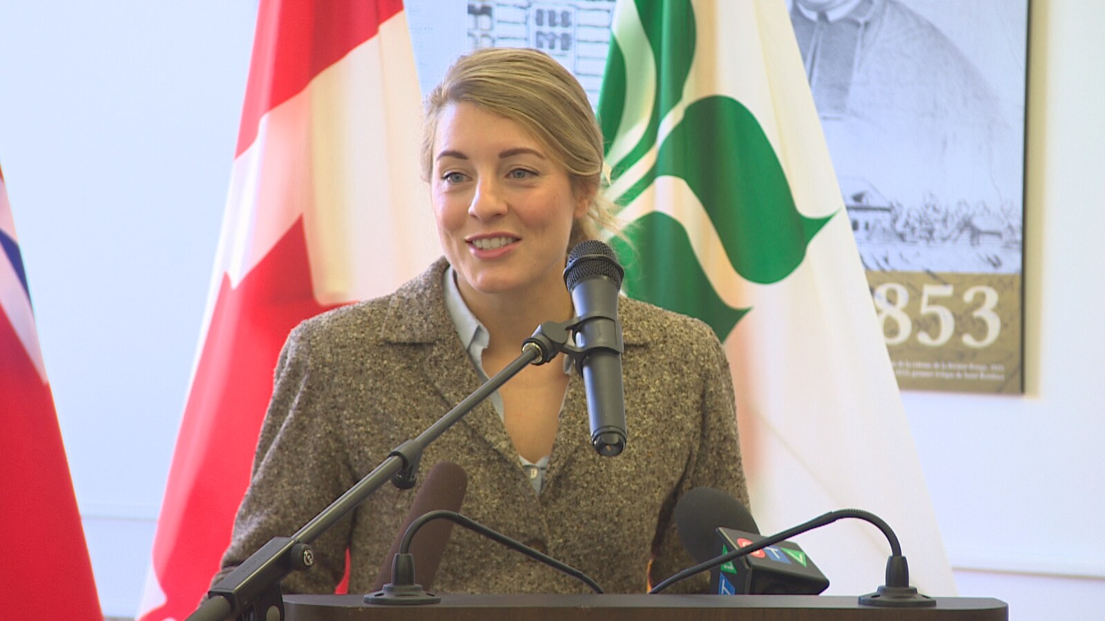 Une femme au micro. Derrière elle le drapeau franco-manitobain ainsi que celui du Canada.
