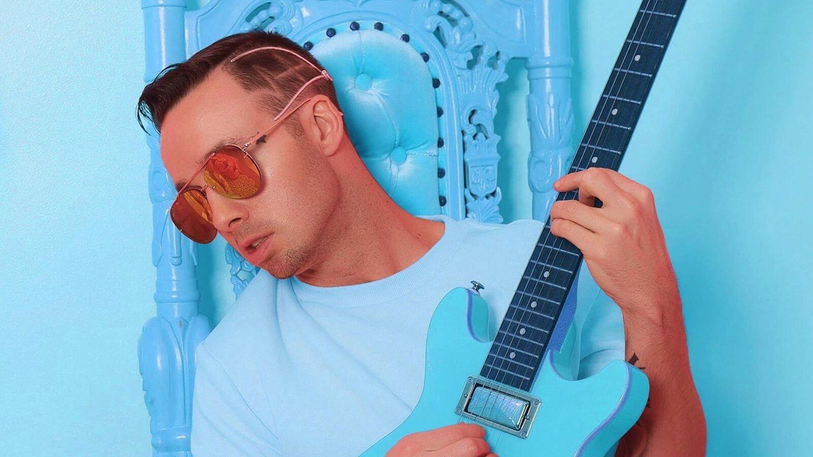 Jon James, vêtu de bleu et de lunettes teintées orange, jouant d'une guitare bleue. Photo promotionnelle.