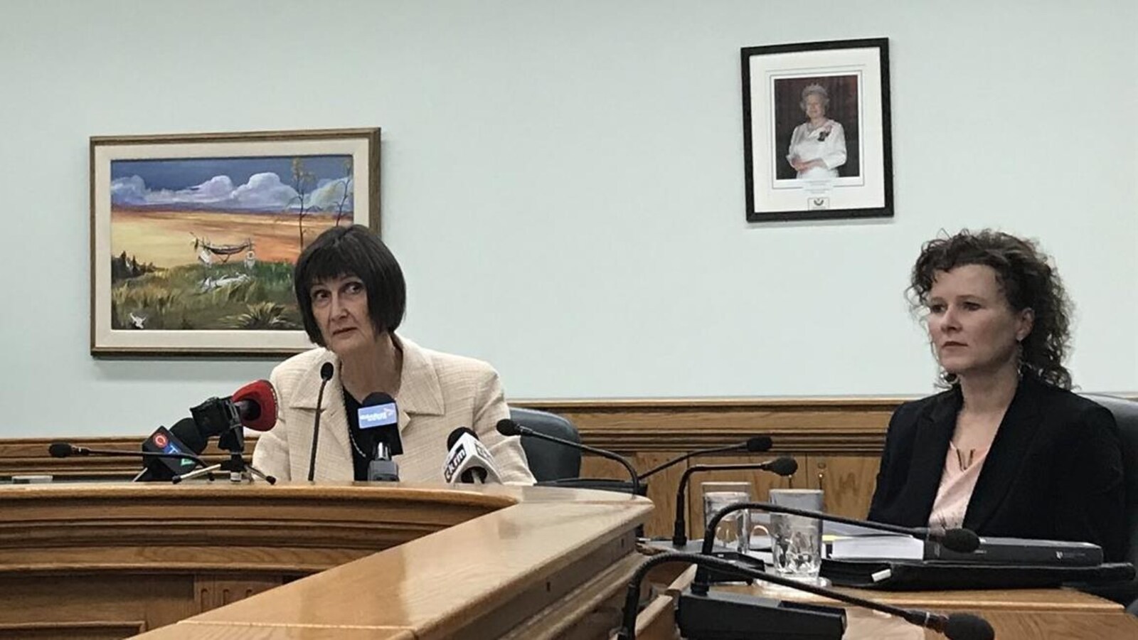 Judy Ferguson parle au micro entourée des membres de son équipe, autour d'un pupitre en bois. Un portrait de la reine est accroché au mur.