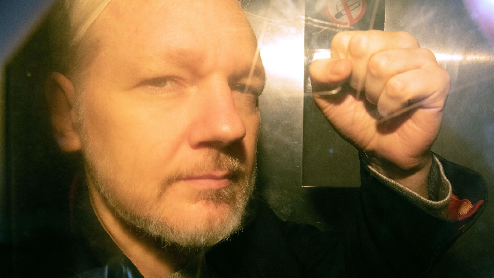 Julien Assange, vu derrière une fenêtre, brandit un poing.