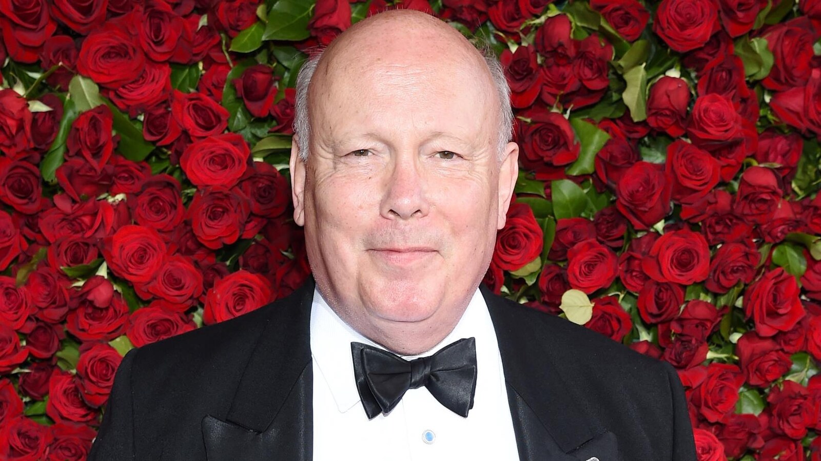 Julian Fellowes vêtu d'un complet noir posant devant un mur de roses rouges.