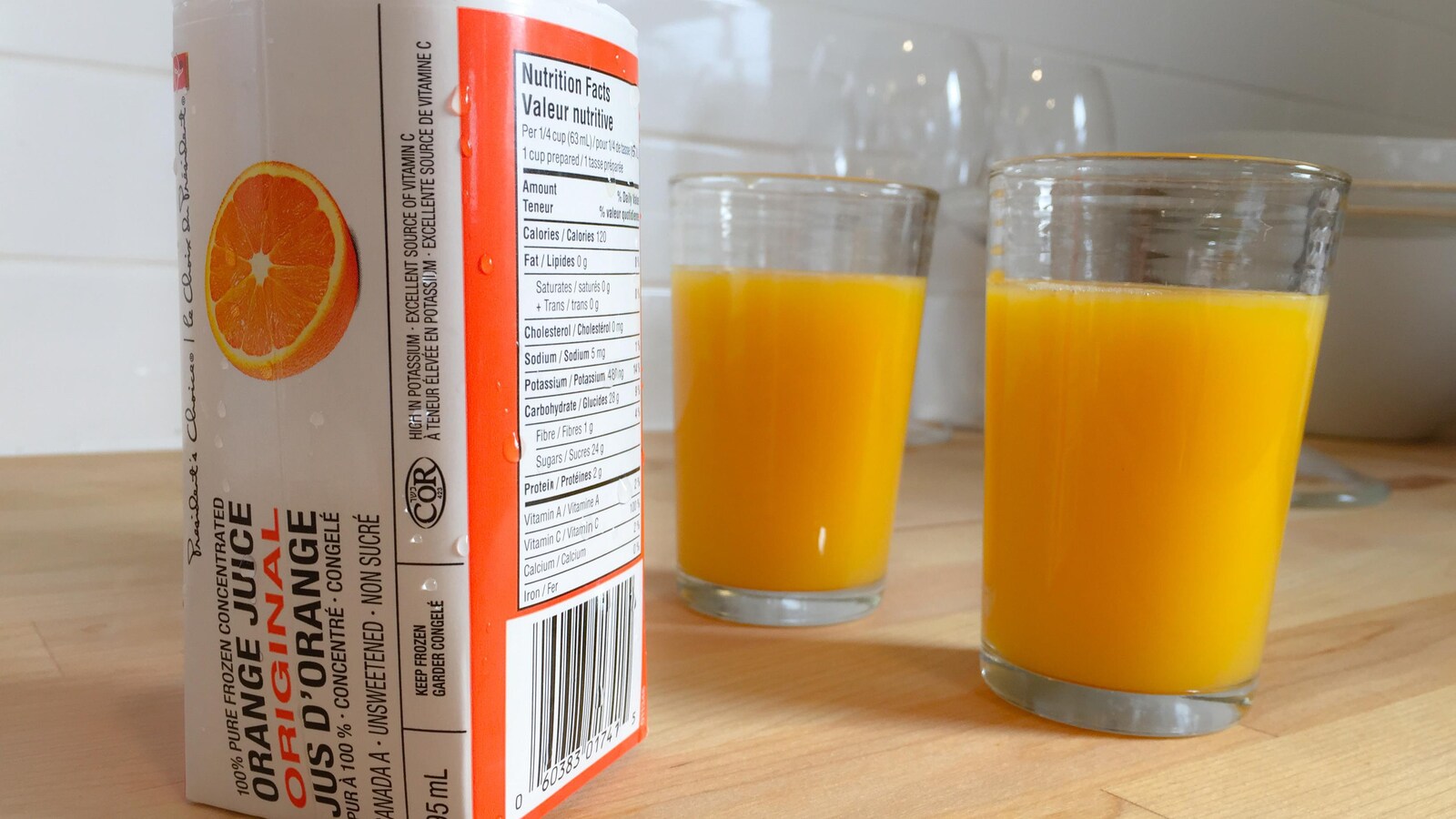 Image d'un contenant de jus d'orange congelé, ainsi que deux verres pleins, sur le comptoir d'une cuisine.