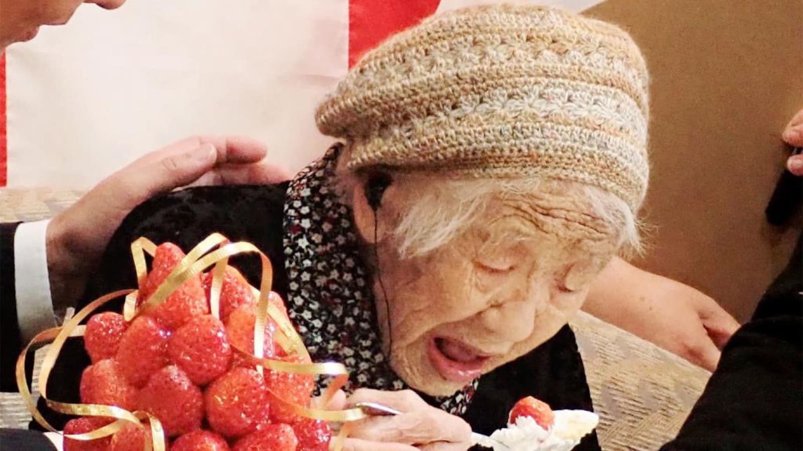 La doyenne de l'humanité, un chapeau sur la tête et une cuillère à la main, se penche devant une assiette, la bouche ouverte, pour manger une part de gâteau à la fraise.