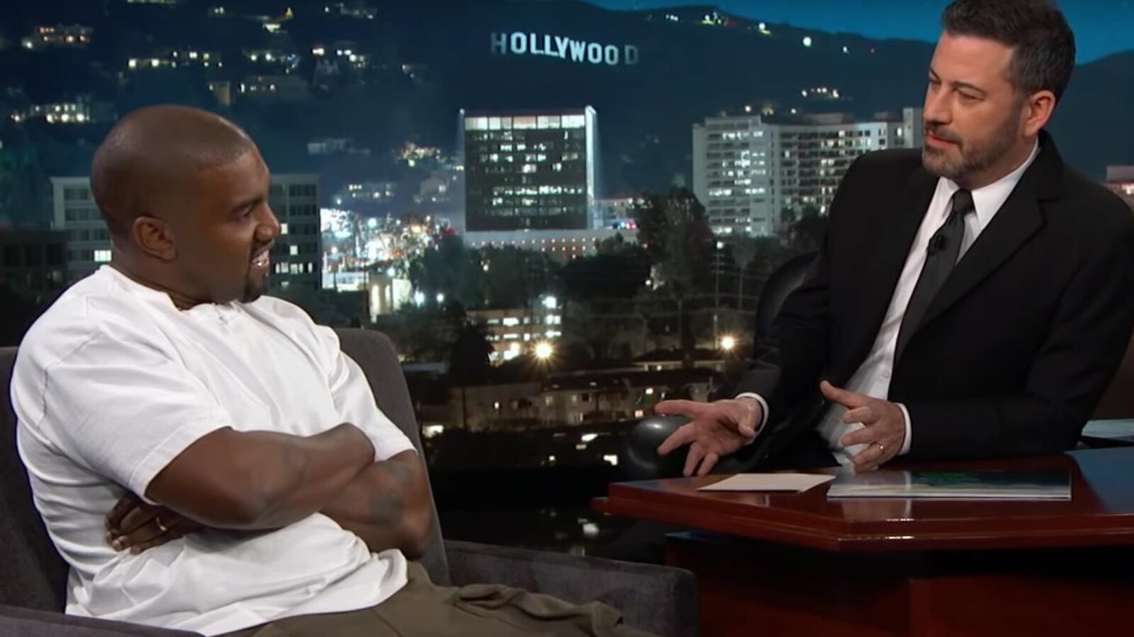 Assis dans un fauteuil, le rappeur Kanye West croise les bras et sourit tandis que l'animateur Jimmy Kimmel, lui aussi assis, lui pose une question.