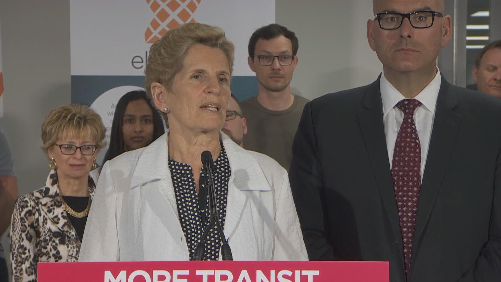 Photo de Kathleen Wynne portant un tailleur blanc, debout derrière un lutrin.