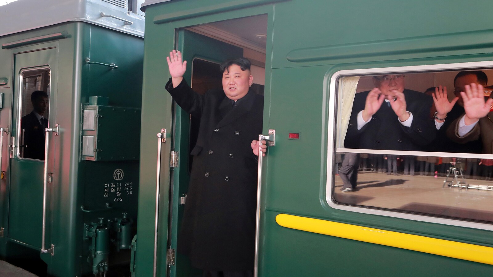 Un homme portant un manteau noir agite la main abord d'un train alors que d'autres hommes font de même à l'intérieur du wagon.
