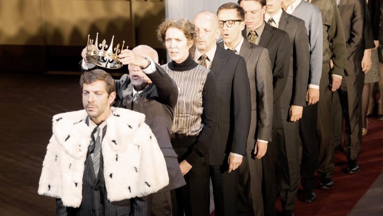 Un homme couronne un autre homme devant lui, tandis qu'une dizaine de personnes fait la file derrière lui, dans une scène de la pièce «Kings of War», du Belge Ivo van Hove.