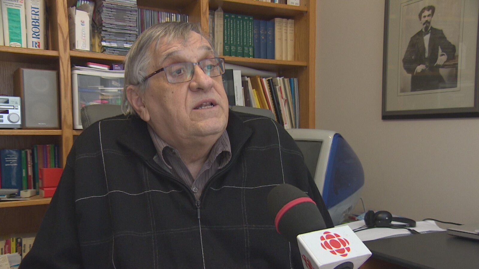L'homme est assis devant une bibliothèque pleine de livres et s'adresse à un micro de Radio-Canada.