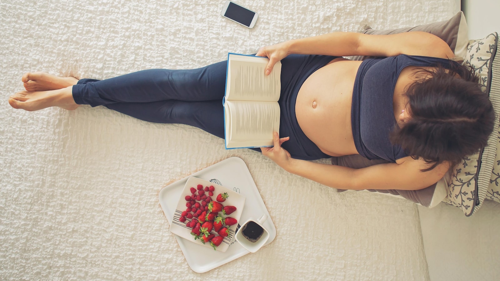 Une femme enceinte lit un livre allongée sur un lit, des fraises à côté d'elle.