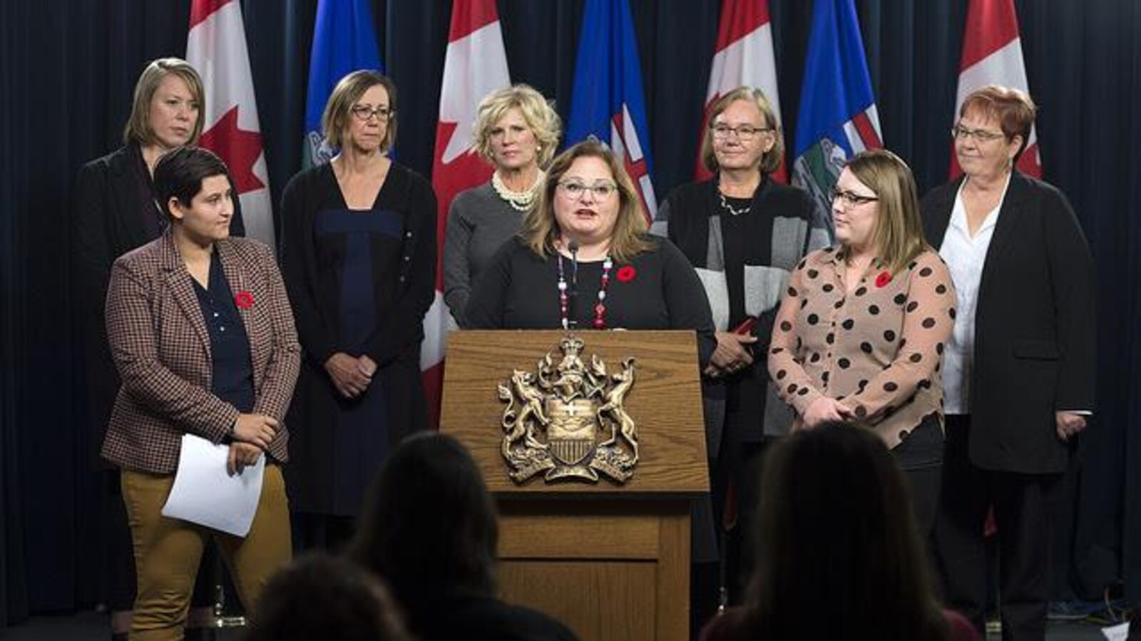 La ministre de la Santé fait un discours. Elle est entourée d'autres femmes. Elle porte des lunettes vertes.