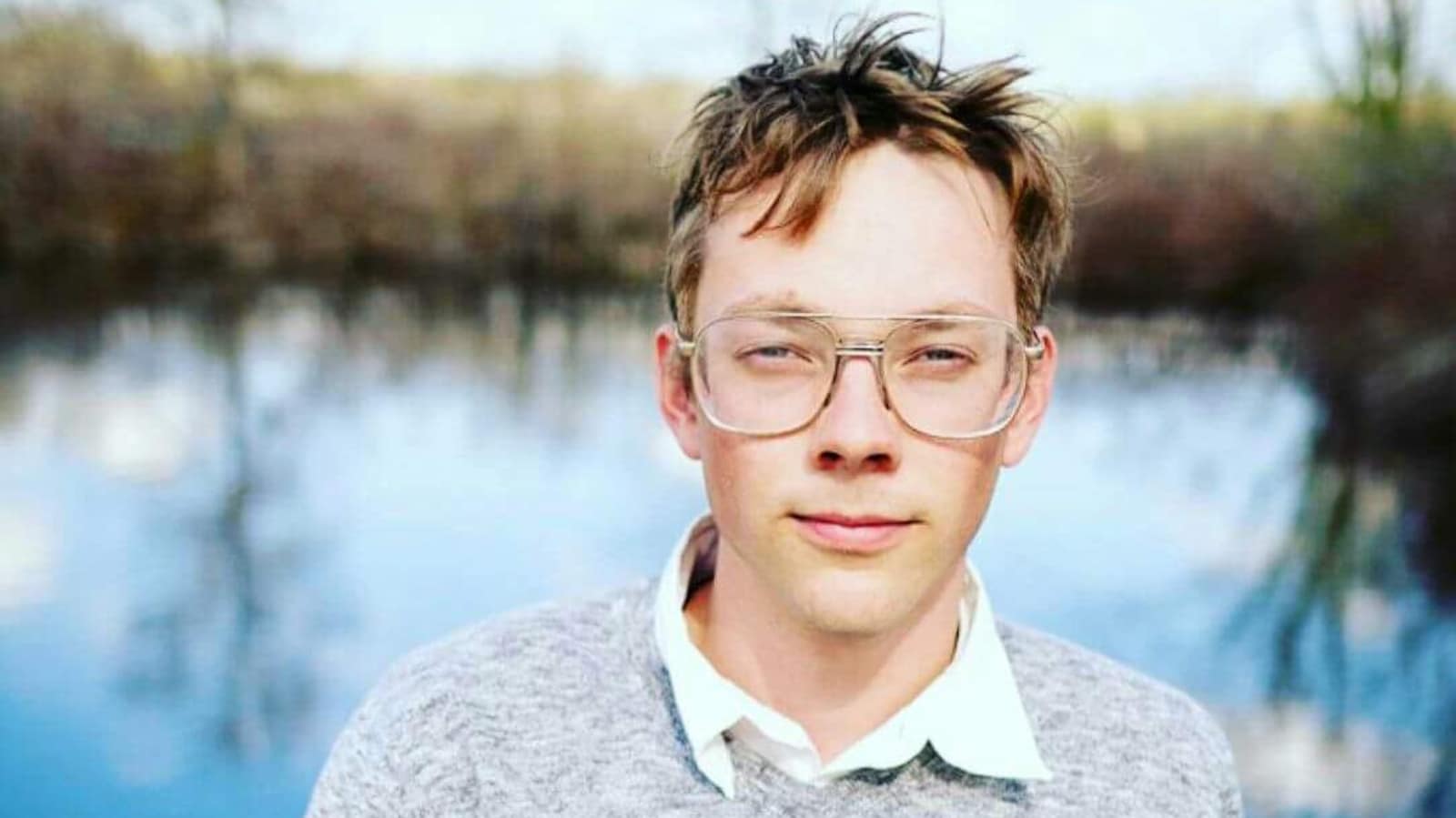 Un jeune homme devant un plan d'eau, aux cheveux courts ébouriffés et aux grosses lunettes vintages, regarde l'objectif.