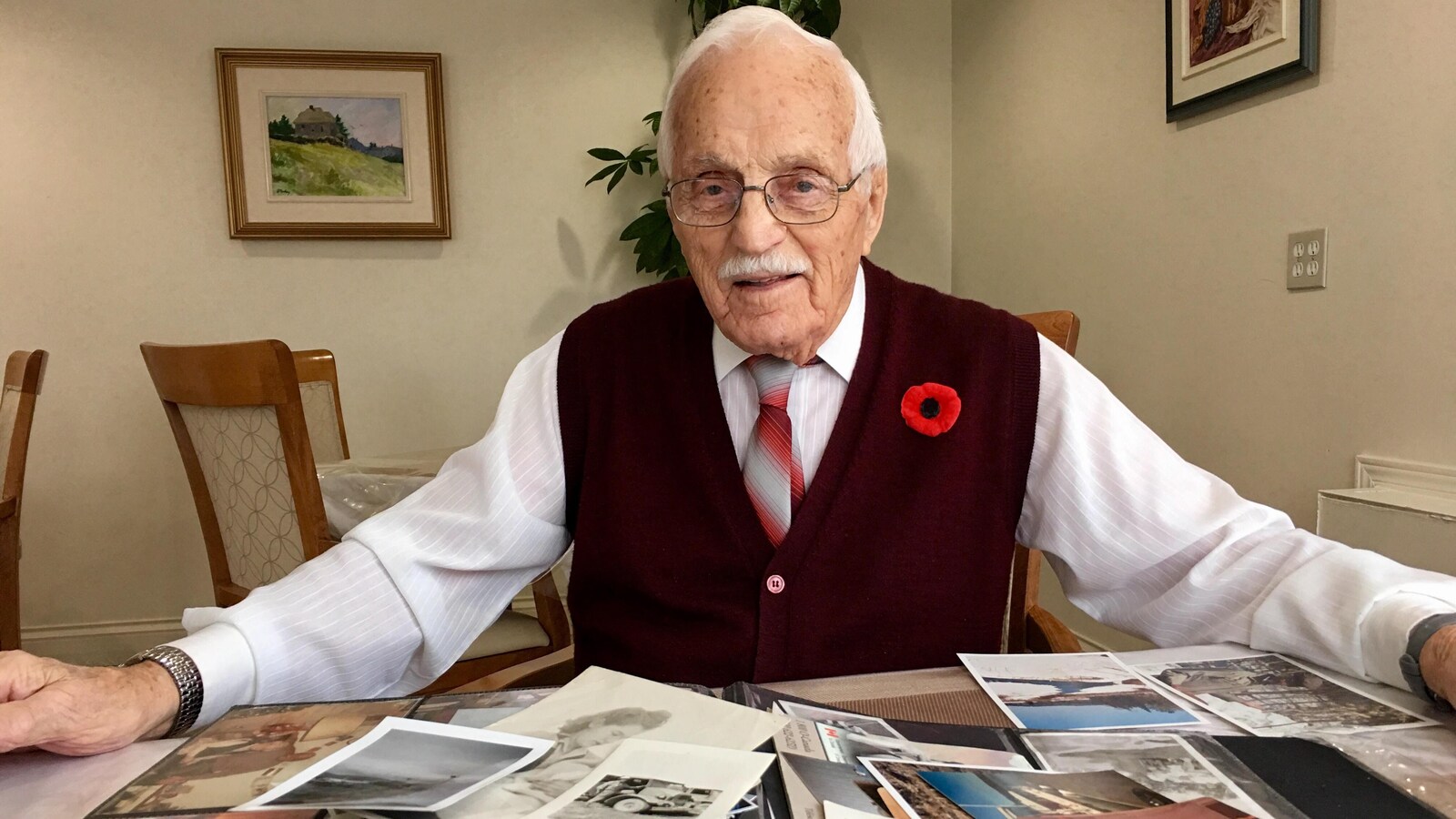 Un homme qui porte un coquelicot à sa veste devant des photos posées pêle-mêle sur un table.