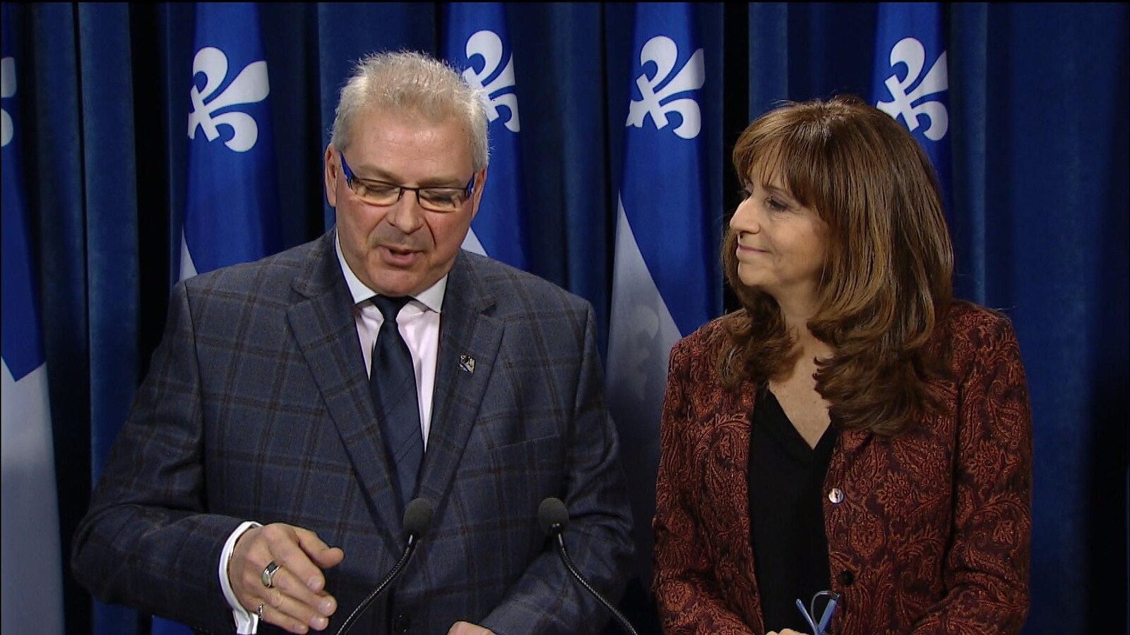 Un homme et une femme debout devant une rangée de drapeaux du Québec.