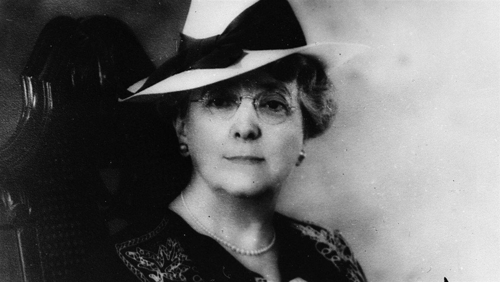 Assise sur une chaise, Lucy Maud Montgomery, portant un chapeau blanc et des lunettes, regarde l'objectif.