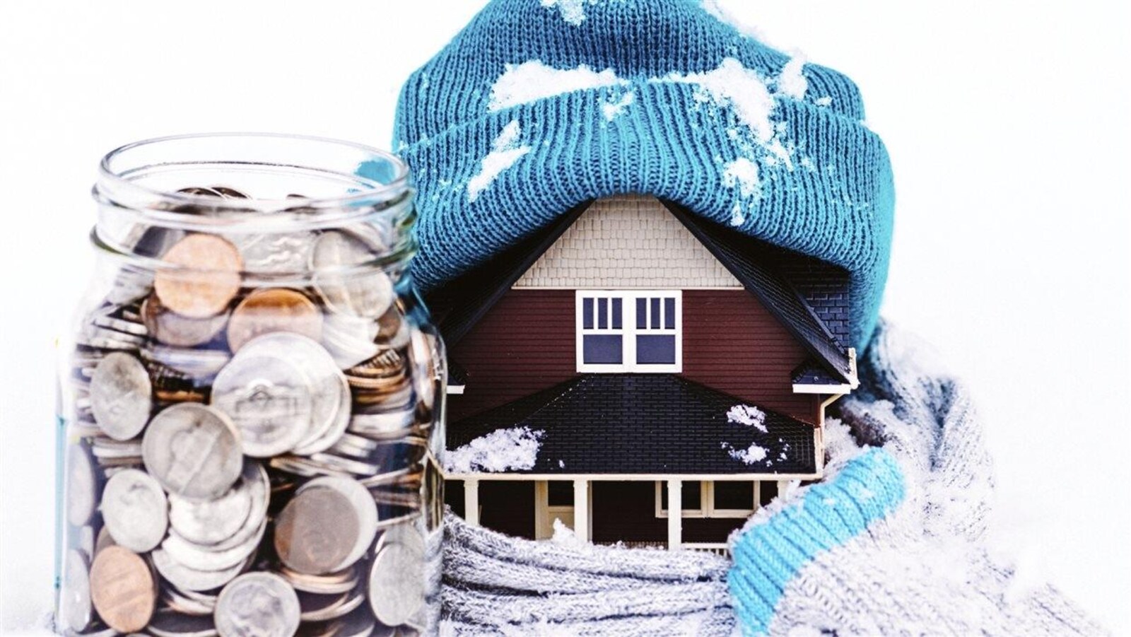 Une maison miniaturisée porte une tuque et un foulard en tenant près d'elle un contenant plein de pièces de monnaie.