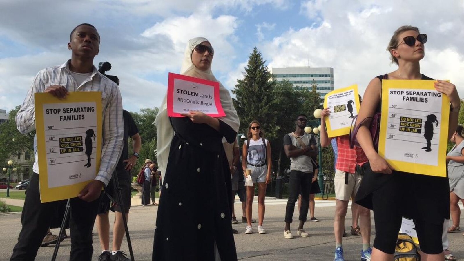 A l'avant-plan, trois personnes tiennent des affiches jaunes et rouges demandant d'arrêter de séparer les familles et contenant le hashtag personne n'est illégal.