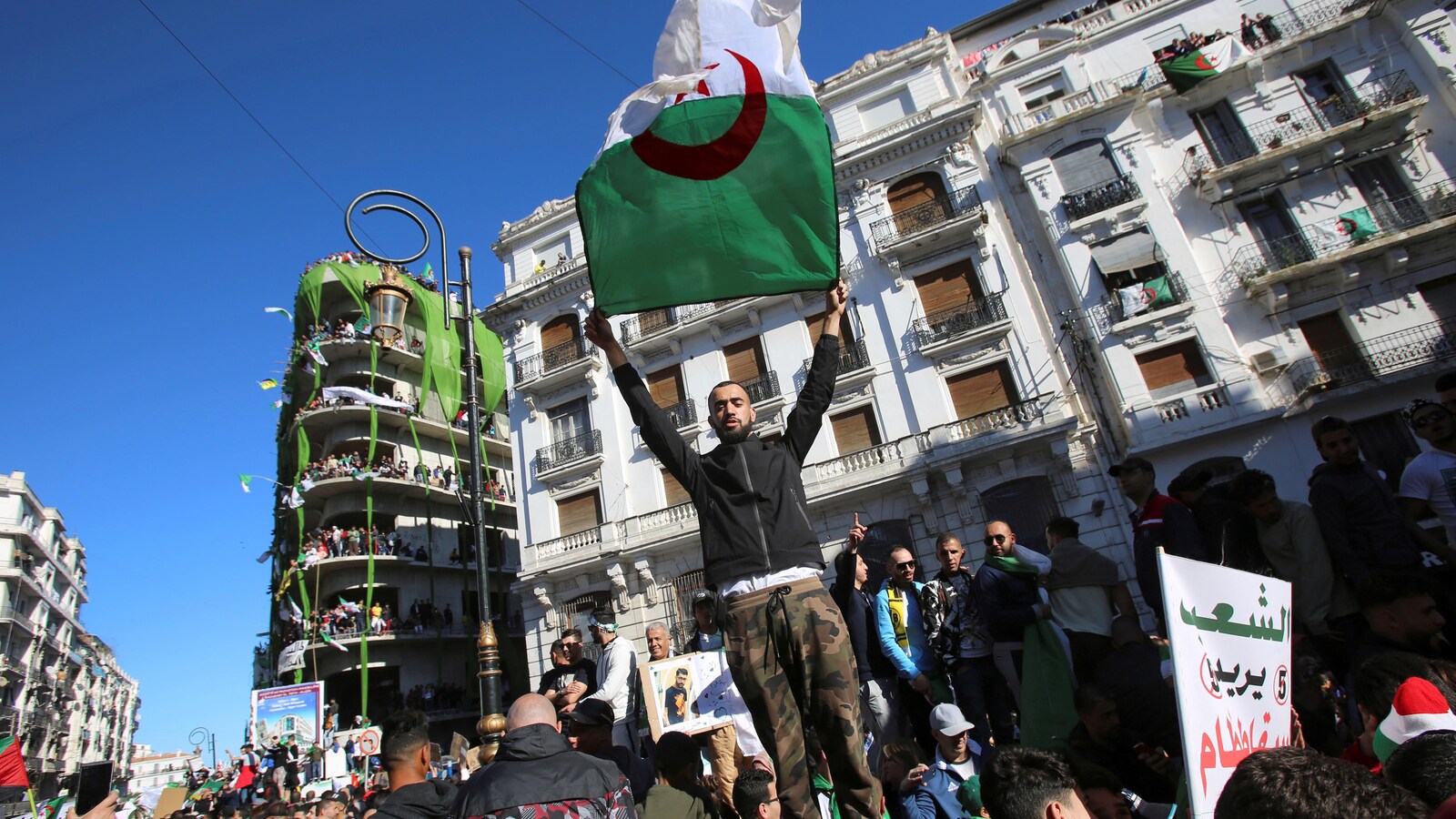 Un homme vêtu d'un pantalon militaire et d'un chandail noir est debout sur un promontoir. Il tend le drapeau algérien, vert à sa gauche, blanc à droite, arborant un croissant rouge et une étoile.