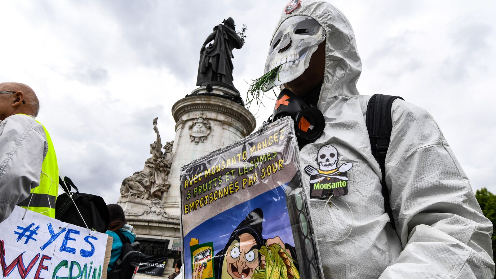 Un homme portant un masque de squelette, une combinaison de protection et des gants tient une pancarte où on peut lire : «Avec Monsanto mangez cinq fruits et légumes empoisonnés par jour».